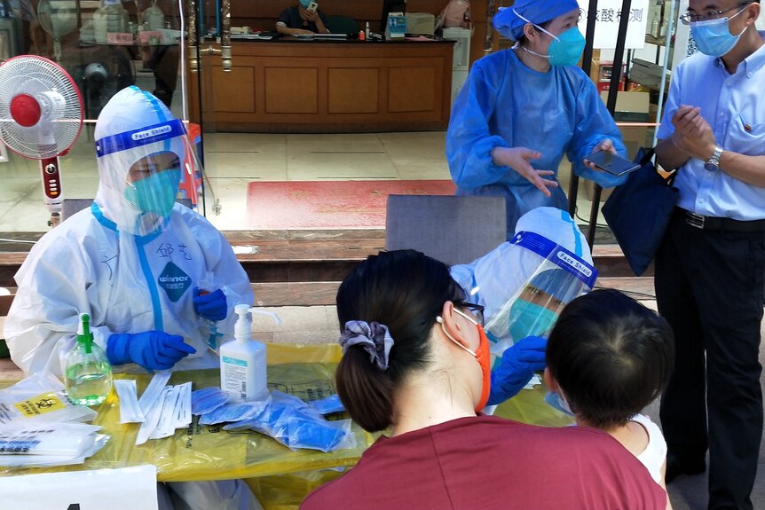 A woman with a face mask holds a child while a medical technician in full PPE swabs the kid's mouth at a covid testing facility.