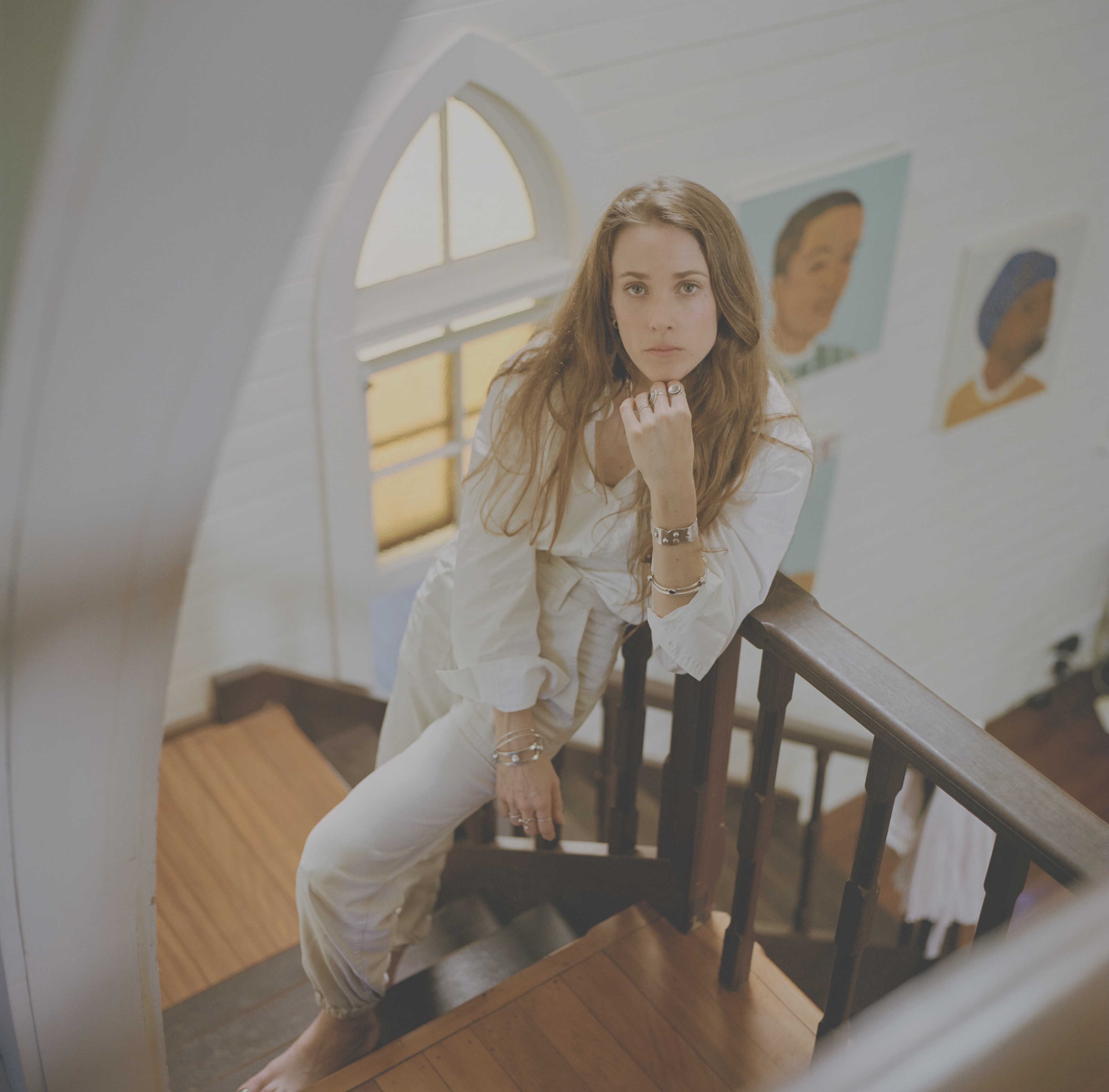 White woman in white shirt & pants with long light brown hair poses on a stairwell, one hand under chin.