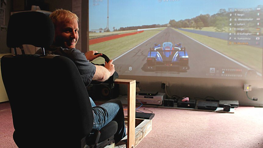 A man sits in a car seat holding a steering wheel with a racetrack on a projector screen.