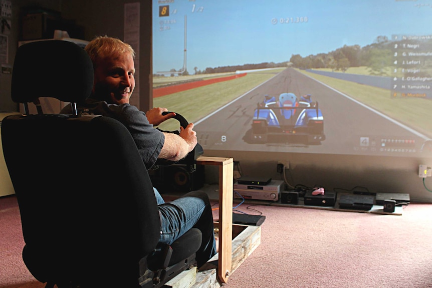 A man sits in a car seat holding a steering wheel with a racetrack on a projector screen.