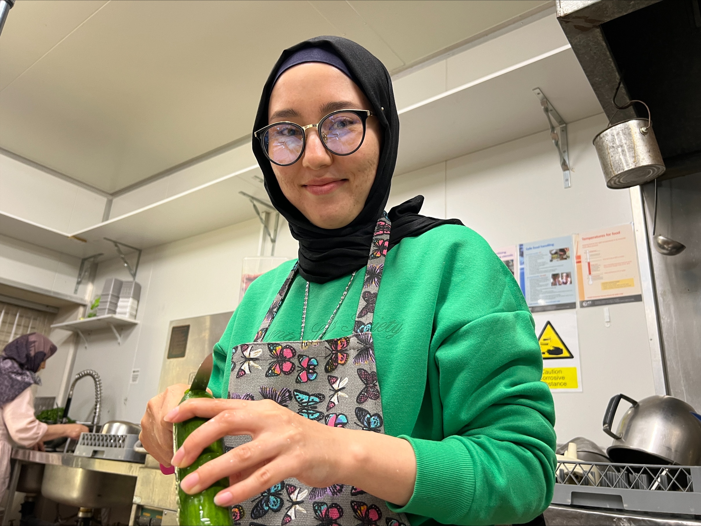 A woman wearing a hijab and glasses smiles in a kitchen as she cuts a cucumber