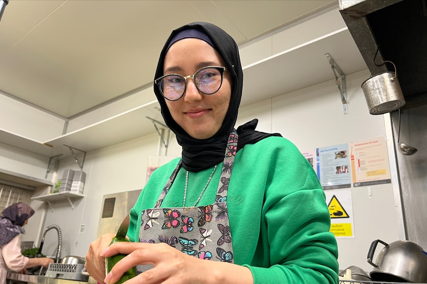 A woman wearing a hijab and glasses smiles in a kitchen as she cuts a cucumber