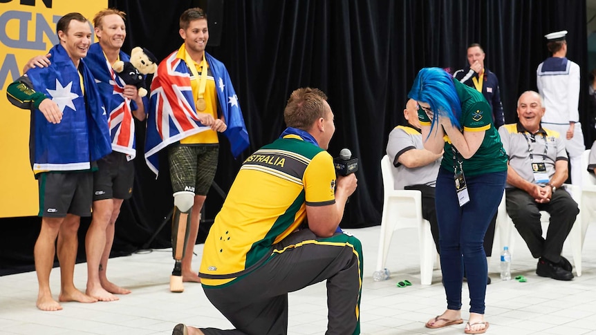 with a backdrop of medal ceremony a man goes down on his knee as a woman holds his face in her hands, bending down in joy