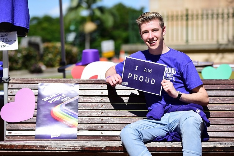 Brock Galway holds up an 'I am proud' sign while sitting on a park bench