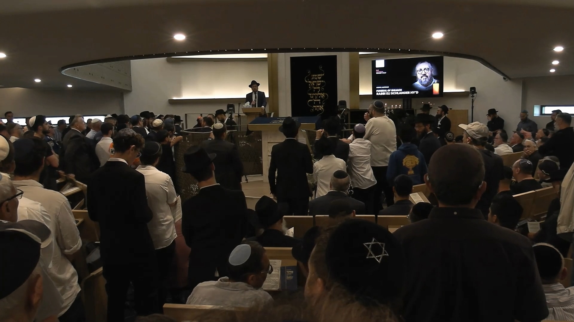 Una gran sala en una sinagoga con muchos dolientes vestidos con kipá y un rabino parado al frente junto a una pantalla.
