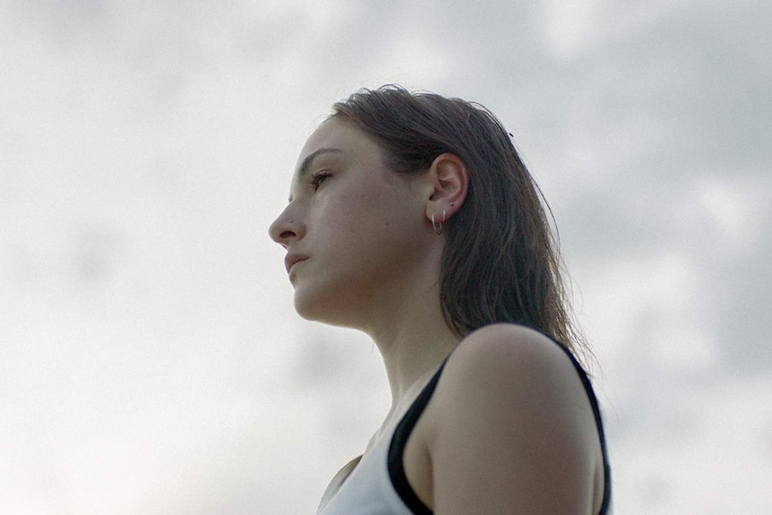 A woman in her early 20s with shoulder length brown hair stares into the distance against a grey clouded sky.