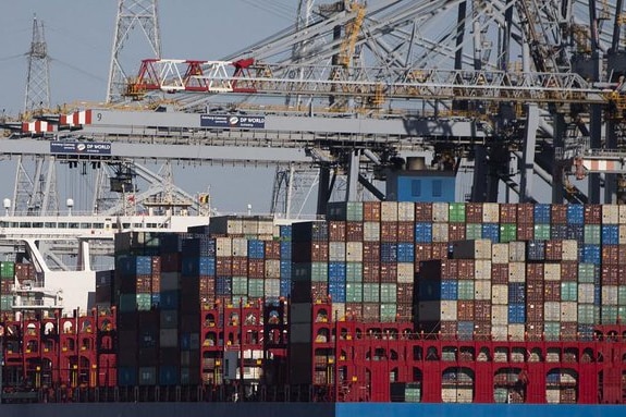 Cargo is lined up at a port with a container ship and cranes behind.