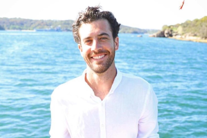 A man wearing a white shirt smiles at the camera with the ocean behind him on a bright sunny day.