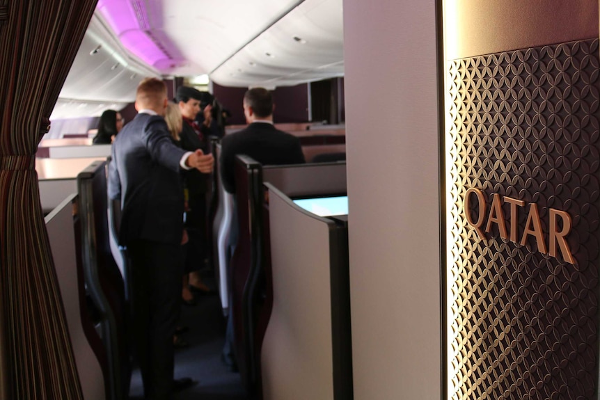 Qatar is written on a wall inside a plane, with media blurred in the background.