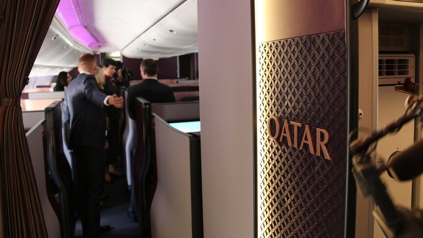 Qatar is written on a wall inside a plane, with media blurred in the background.