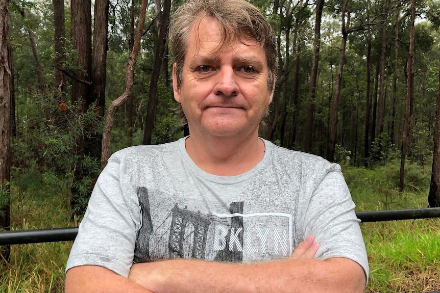 A man stands with his arms crossed in the bush with trees behind him