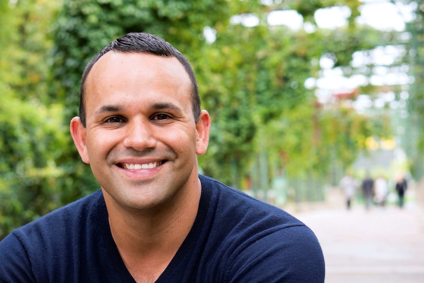 A head and shoulders image of a young man smiling against a green leafy outdoor backdrop.