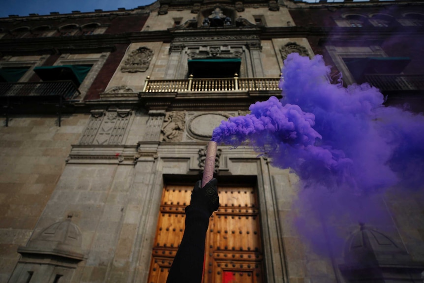A gloved hand holds up a candle with purple smoke billowing out of it in front of Mexico's National Palace.