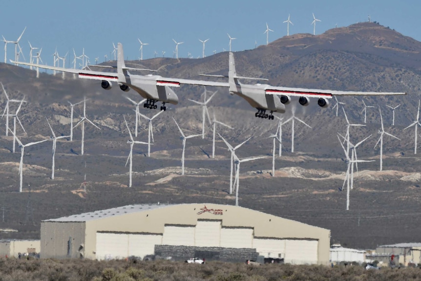 A large six-engine aircraft flies in front of wind farms.