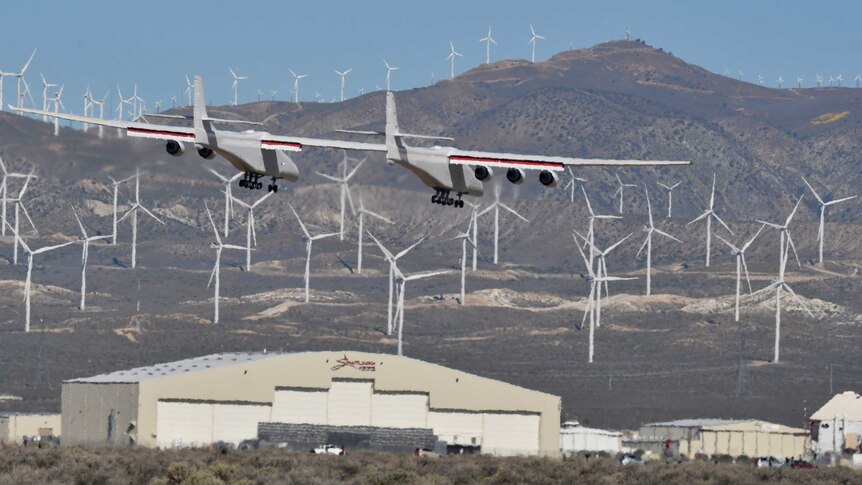 A large six-engine aircraft flies in front of wind farms.