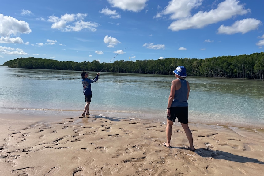 Two people stand on a sandy beach. The boy is holding a fishing rod. 