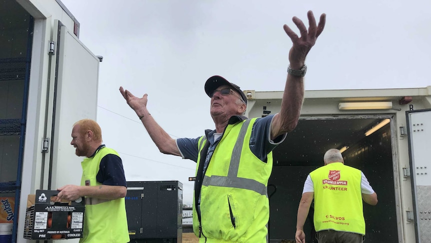 A man in a yellow hi-vis vest throws his hands up in the air as it rains around him