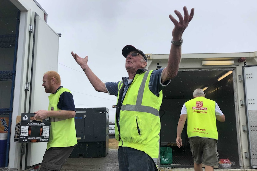 A man in a yellow hi-vis vest throws his hands up in the air as it rains around him