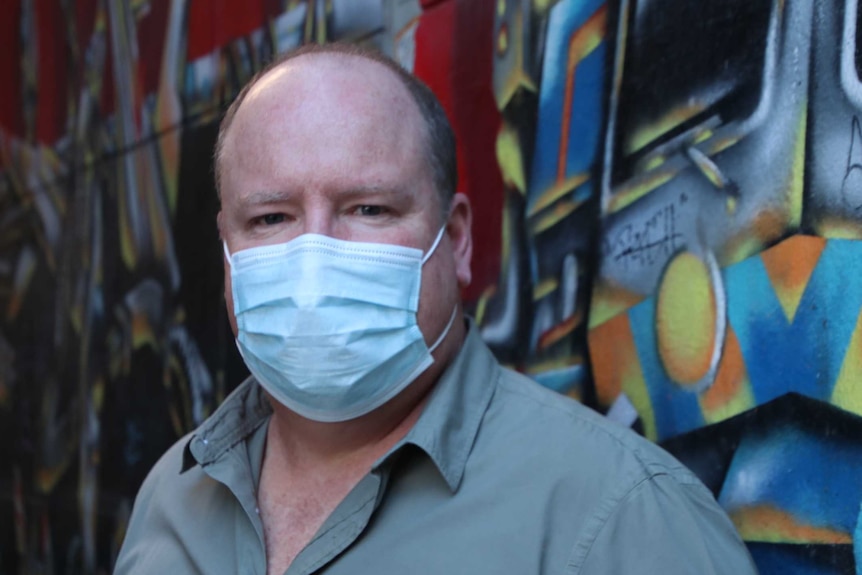 A man wearing a face mask stands in an alley in front of a wall of graffiti.