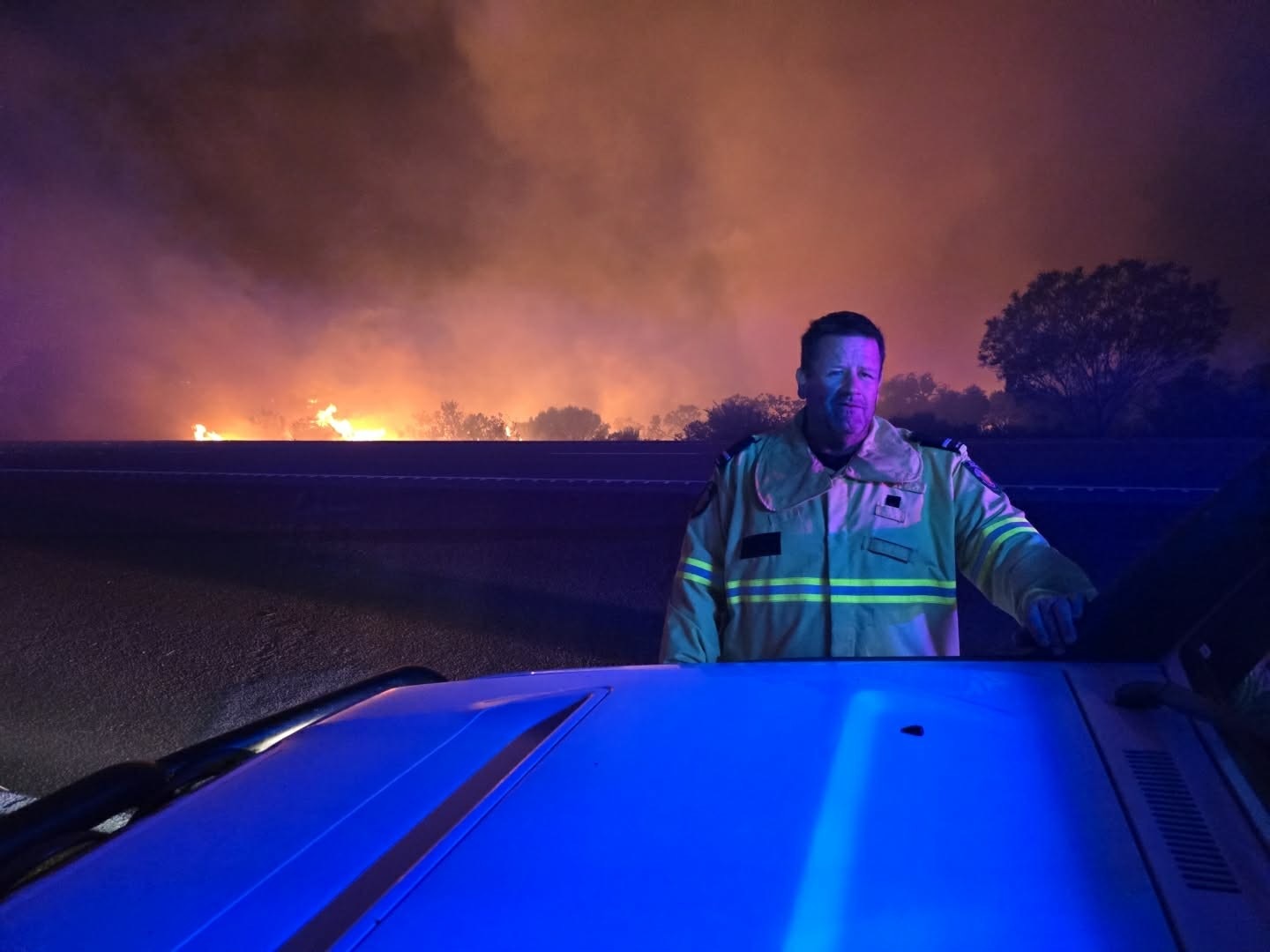 Um homem parado ao lado de um veículo com um incêndio florestal ao fundo