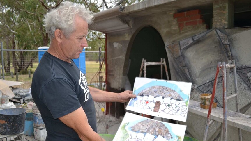 The artists stands in front of the construction site holding two hand drawn impressions of the final result.