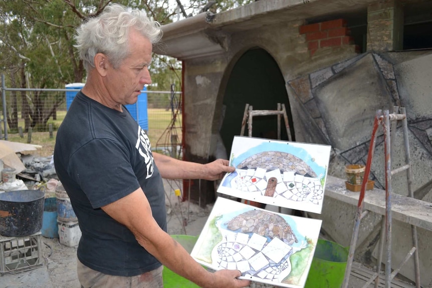 The artists stands in front of the construction site holding two hand drawn impressions of the final result.