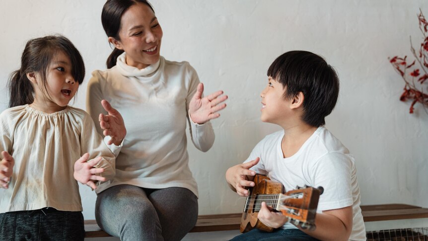 A young boy playing ukulele for a younger girl and an adult woman