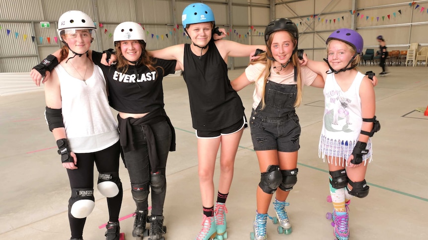 Five girls with helmets and skates on pose for a photo in a huge warehouse.