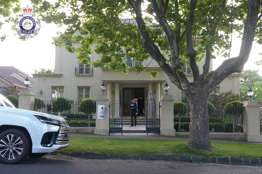A police officer in front of a large double-storey home on a leafy street.