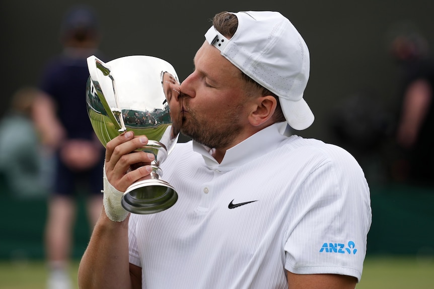 A wheelchair tennis player in white shirt and cap kisses a large silver trophy cup
