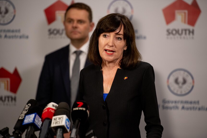 A woman in black blazer speaking into microphones. A man stands behind her in front of South Australian government banner