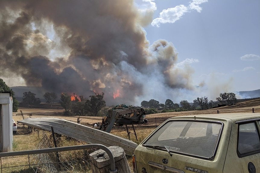 An old station wagon sits in the foreground, as smoke billows above roaring flames in the neighbouring paddock.