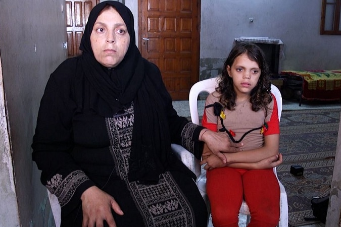 A mother places her hand on her sick daughter's lap as they sit on white plastic chairs in the doorway of a home.