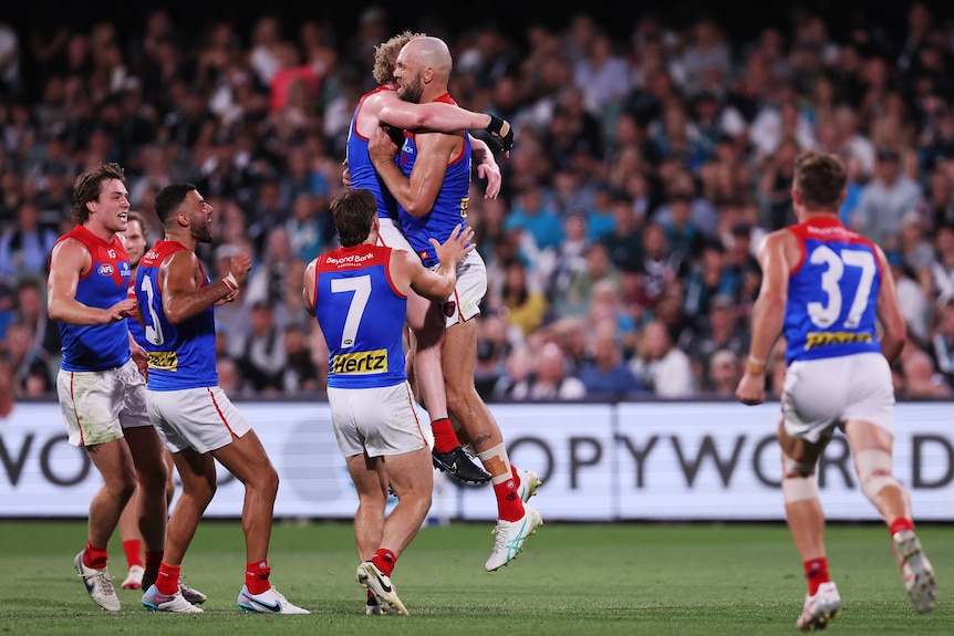 Max Gawn dari Melbourne berpelukan di udara dengan rekan setimnya saat timnya berlari untuk memberi selamat kepadanya setelah mencetak gol. 