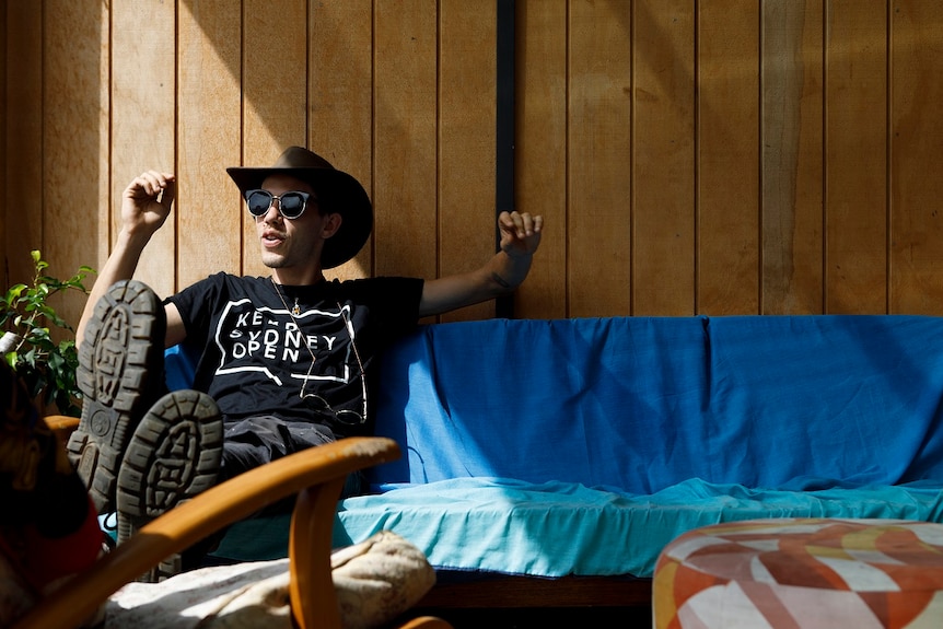A man sits on a seat in the sun wearing a hat and sunglasses and with his boots up on a chair for a story on share housing.