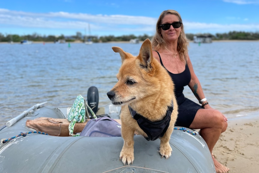 a woman and her dog sitting on a small boat on the bank of a bay