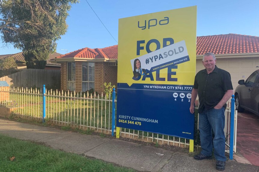 A man smiles as he stands in front of a for sale sign at his house in the outer Melbourne suburbs.