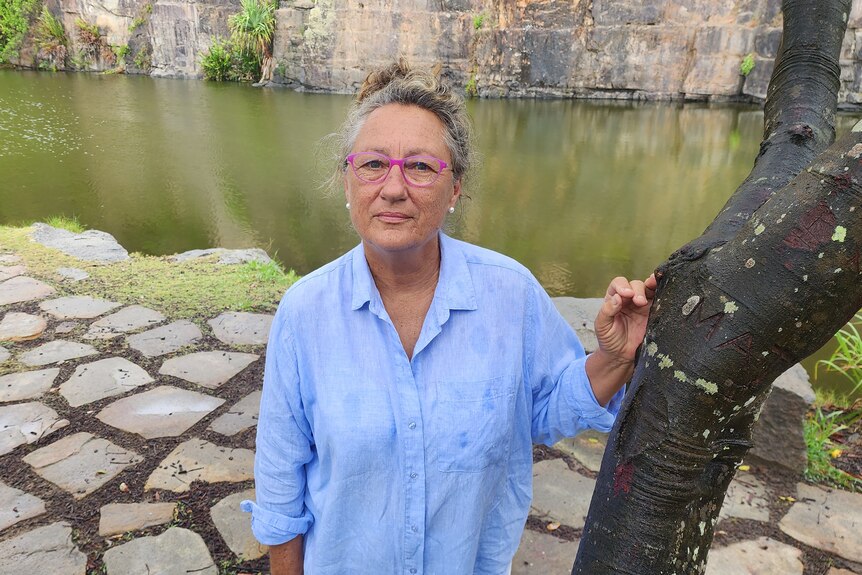A middle-aged, bespectacled woman leans against a tree in front of a waterhole.