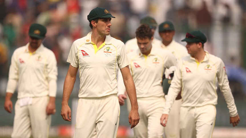 Pat Cummins walks in front of a group of cricketers in white clothes