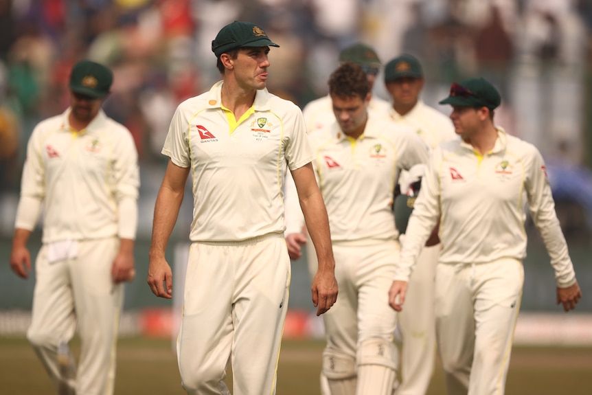 Pat Cummins walks in front of a group of cricketers in white clothes