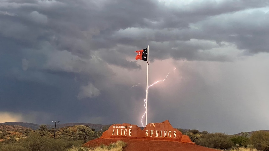 The welcome to Alice Spring sign, with the NT flag, a stormy sky behind it and a bolt of lightning in the background.