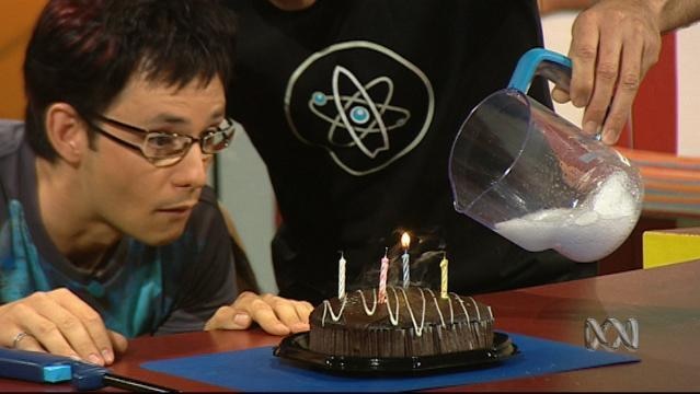 Hand tips jug of white foam above a birthday cake with lit candle as another man watches