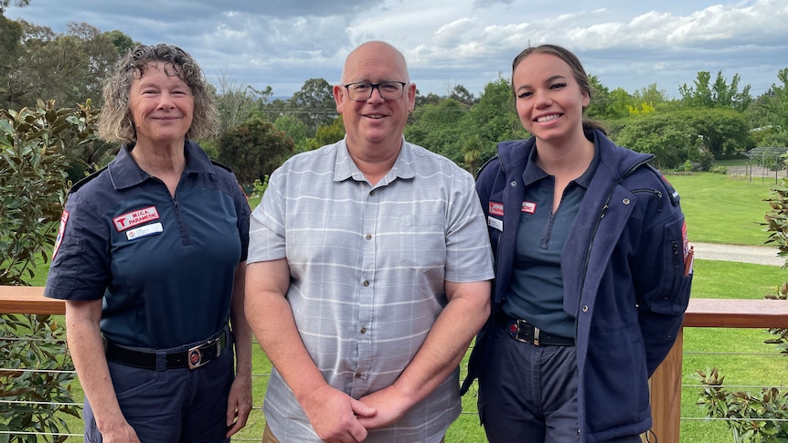 Two uniformed paramedics stand on either side of a smiling, middle-aged man on a rural property beneath a cloud-swept sky.