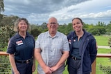 Two uniformed paramedics stand on either side of a smiling, middle-aged man on a rural property beneath a cloud-swept sky.