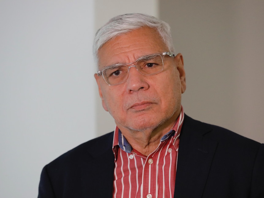 A Frist Nations man, glasses, red shirt with white stripes, dark blazer, looks on with a neutral expression