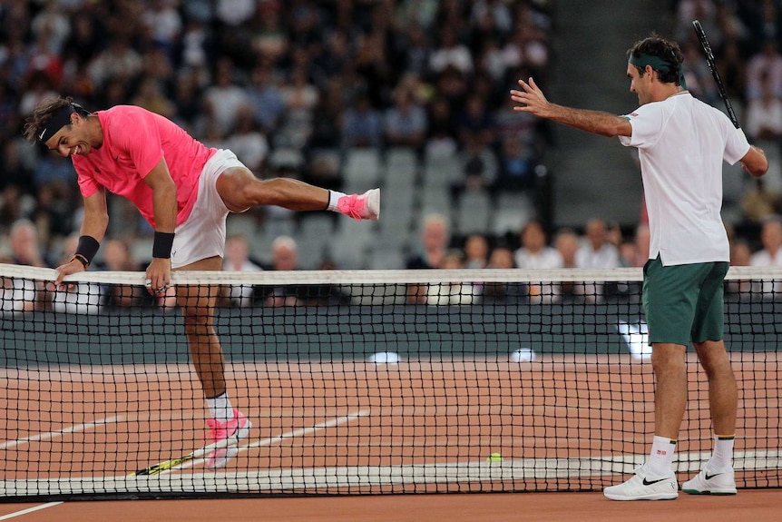 Roger Federer holds his hands out as Rafael Nadal hurdles the net with a big smile on his face