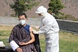 A man in traditional Bhutanese dress receiving an injection from a health worker in full PPE