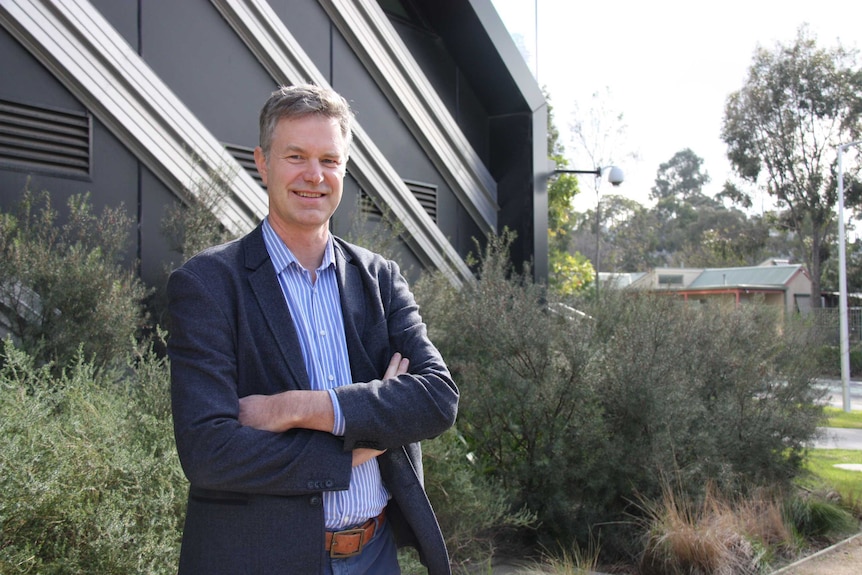 A man in a blue suit and blue shirt stands in front of green bushes and a black building.