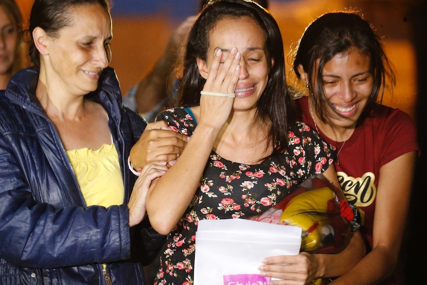 A young woman covered part of her face with one hand and is crying with a smile on her face flanked by her mother and family.