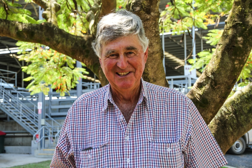 A man with a check shirt stands smiling in front of a large tree at a livestock saleyard.
